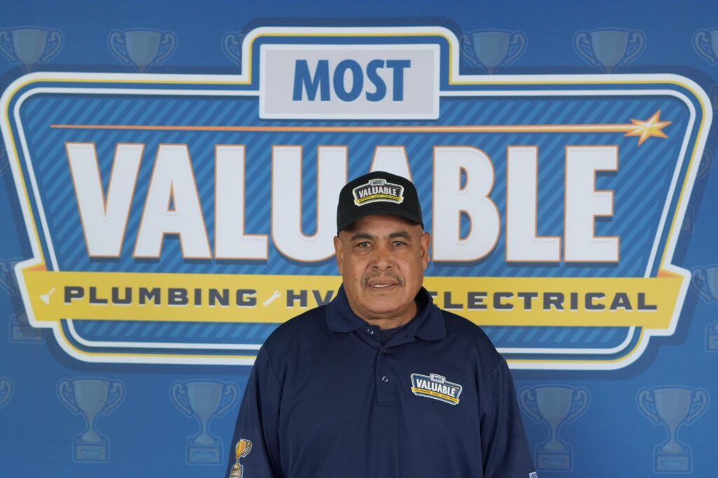 A man wearing a "Most Valuable" cap and shirt stands in front of a sign reading "Most Valuable Plumbing HVAC Electrical," ready to welcome you to meet the team.