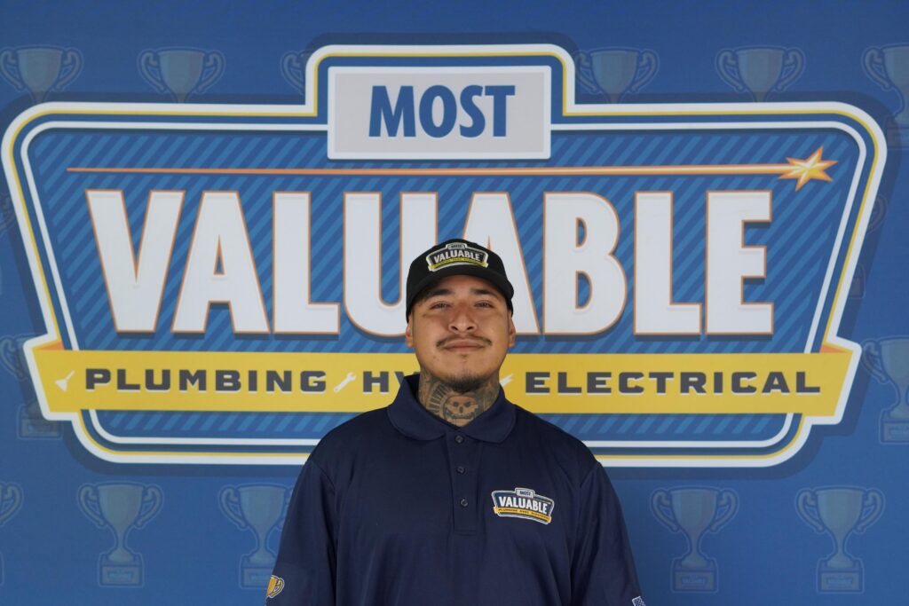 A man in a "Most Valuable" uniform stands in front of a sign that reads "Most Valuable Plumbing • HVAC • Electrical," ready to help you meet the team behind your trusted home services.