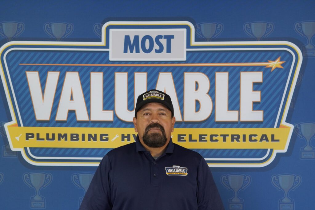 A man in a "Most Valuable" uniform stands in front of a sign that reads "Most Valuable Plumbing HVAC Electrical," with trophy icons in the background—meet the team delivering award-winning service.