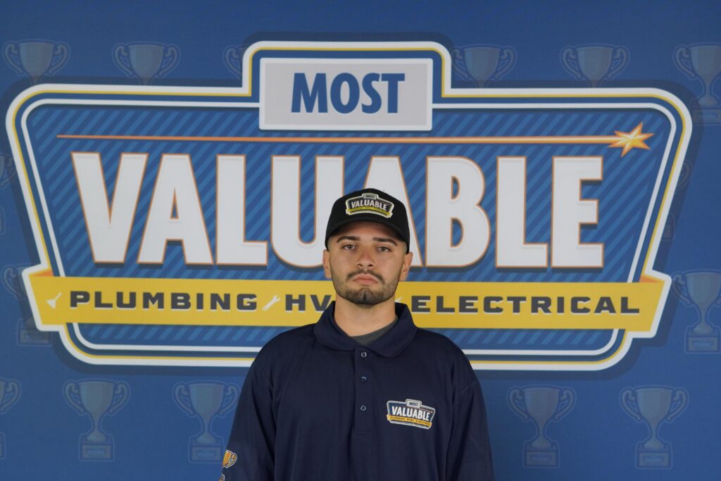 A man in a navy uniform and cap stands in front of a "Most Valuable Plumbing HVAC Electrical" backdrop decorated with trophy icons, ready to meet the team.