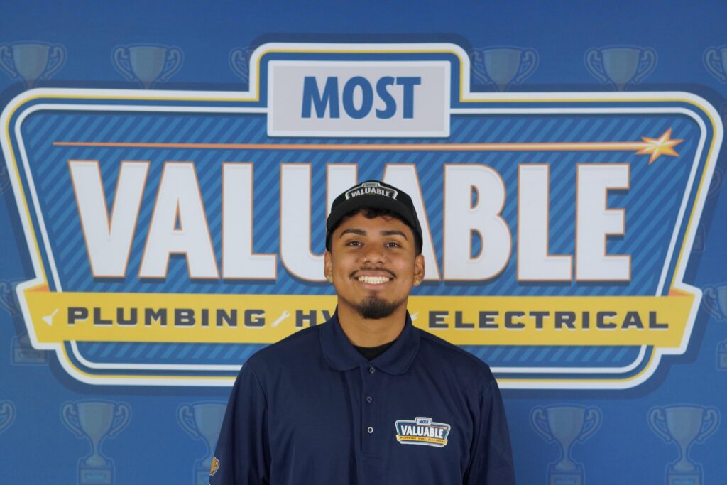 A man in a "Most Valuable" uniform and cap smiles in front of a backdrop that reads "Most Valuable Plumbing HVAC Electrical," ready to welcome you as part of our meet the team spotlight.
