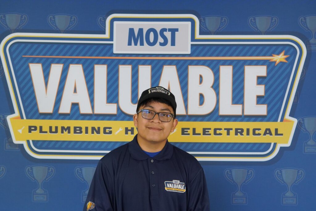 A person wearing glasses and a uniform stands in front of a backdrop that reads "Most Valuable Plumbing • HVAC • Electrical," ready to welcome you as part of the meet the team experience.