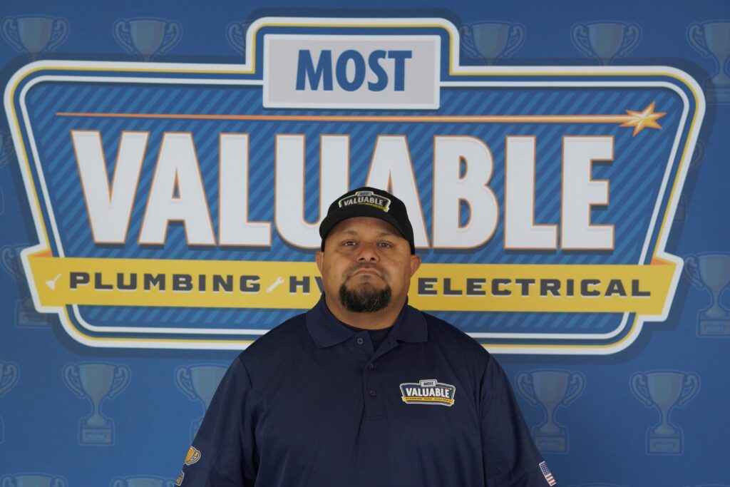 Meet the team: A man in a blue “Most Valuable” uniform stands in front of a sign that reads “Most Valuable Plumbing HVAC Electrical,” set against a trophy pattern background.